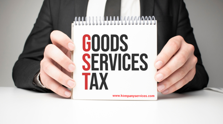 A person in a suit is holding a spiral notebook with the words "Goods Services Tax" written on it. The first letters of each word are red, forming the acronym "GST." Below, the URL www.kompanyservices.com is displayed, highlighting an important decision by the GST Council.