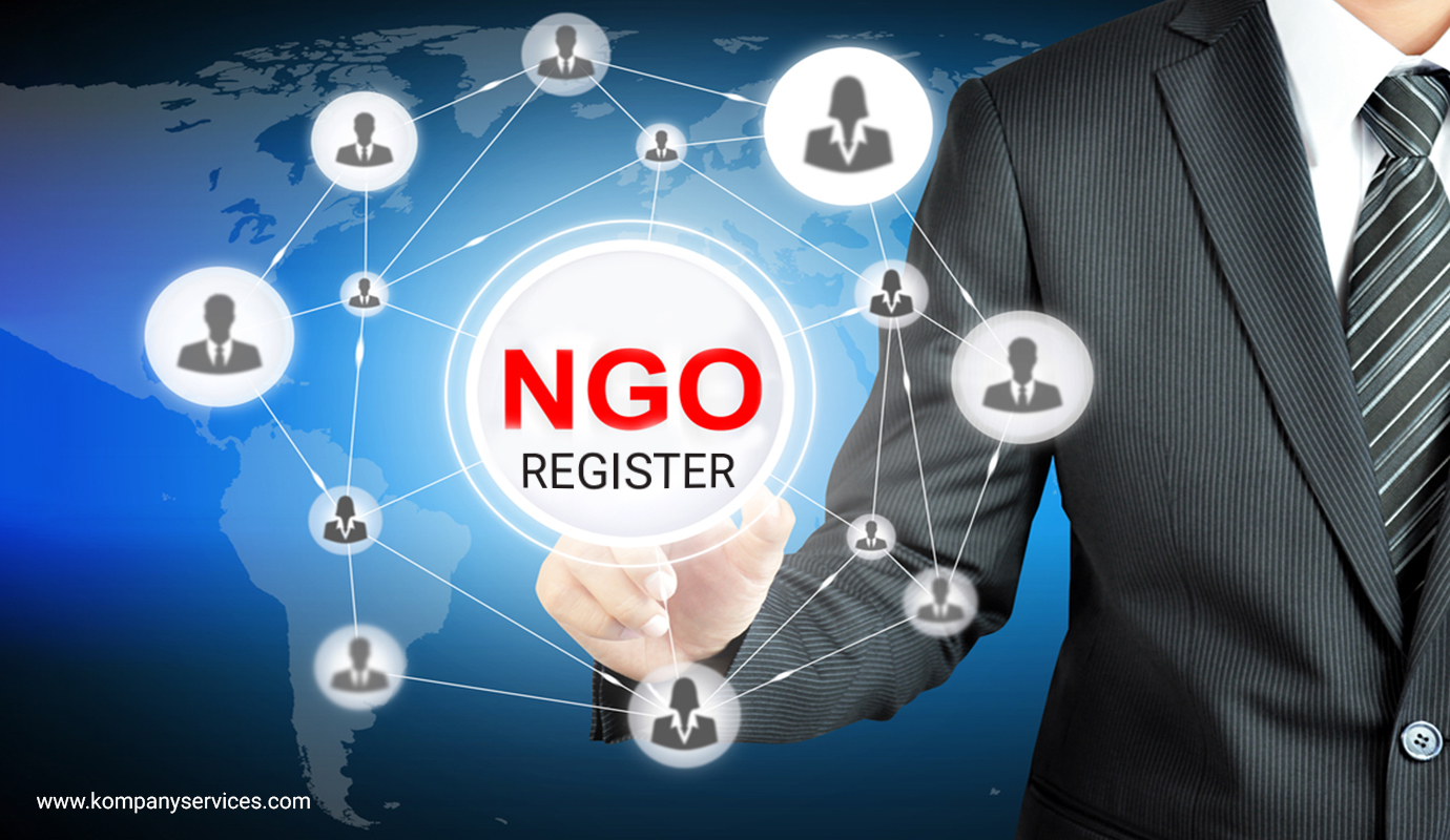 A businessman in a suit points at a digital interface displaying "NGO REGISTER" in red text, symbolizing NGO registration. Surrounding the text are icons of people connected in a network, with a world map backdrop. The URL www.kompanyservices.com is visible at the bottom left corner.