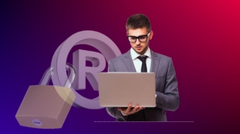 A man wearing glasses and a suit stands looking at a laptop in his hands. In the background, there's a large padlock and a registered trademark symbol, emphasizing the importance of trademark registration. The color gradient of the background is a blend of purple and red.